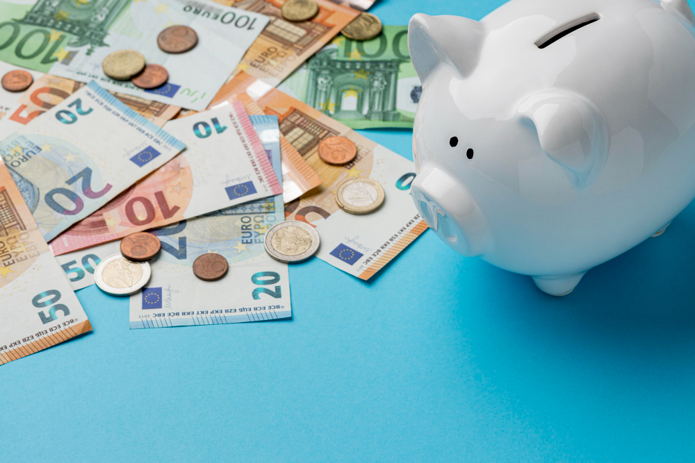 Mehrere Euro Scheine, sowie Münzen liegen auf einem Hellblauen hntergrund neben einem Sparschwein